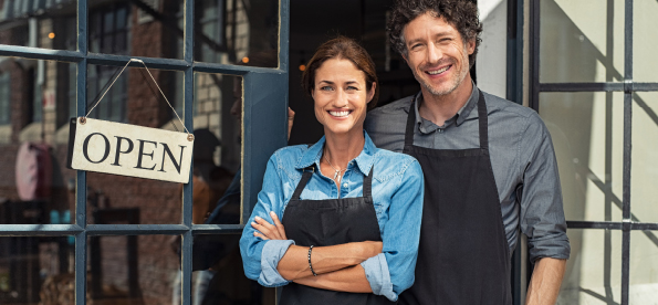 Two cheerful small business owners smiling and looking at camera while standing at entrance door  Happy mature man and mid woman at entrance of newly opened restaurant with open sign board 